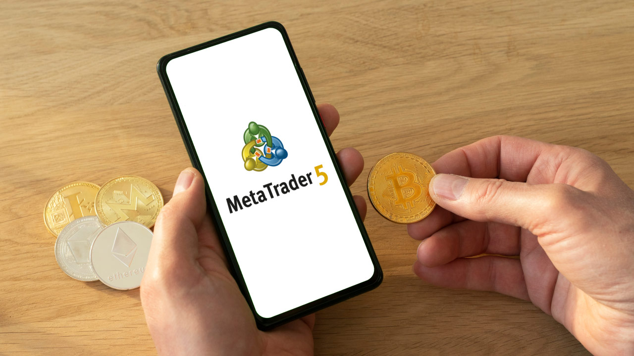 A person holding a Bitcoin coin in one hand while using a smartphone displaying the MetaTrader 5 logo, with various cryptocurrency coins on a wooden table, representing trading cryptocurrencies with MetaTrader 5.
