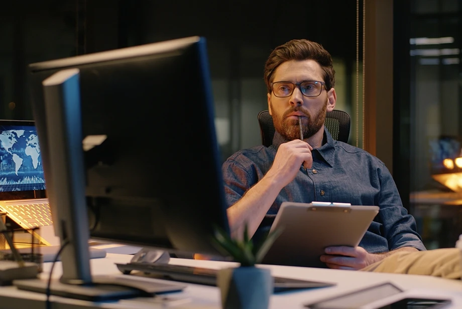 A man sitting at a desk in an office at night, holding a clipboard and pen while thoughtfully looking at a computer monitor.