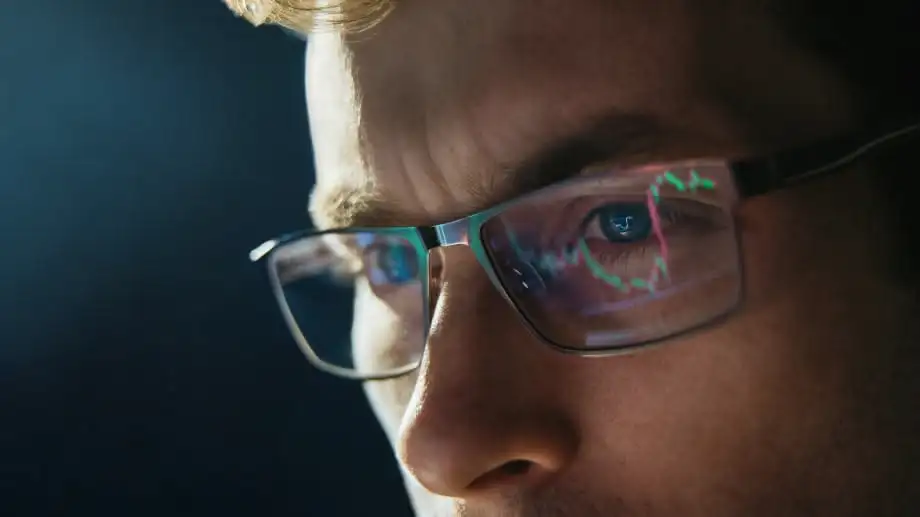 Close-up of a trader’s face with financial charts reflected in their glasses, showing focus and market analysis on a screen.